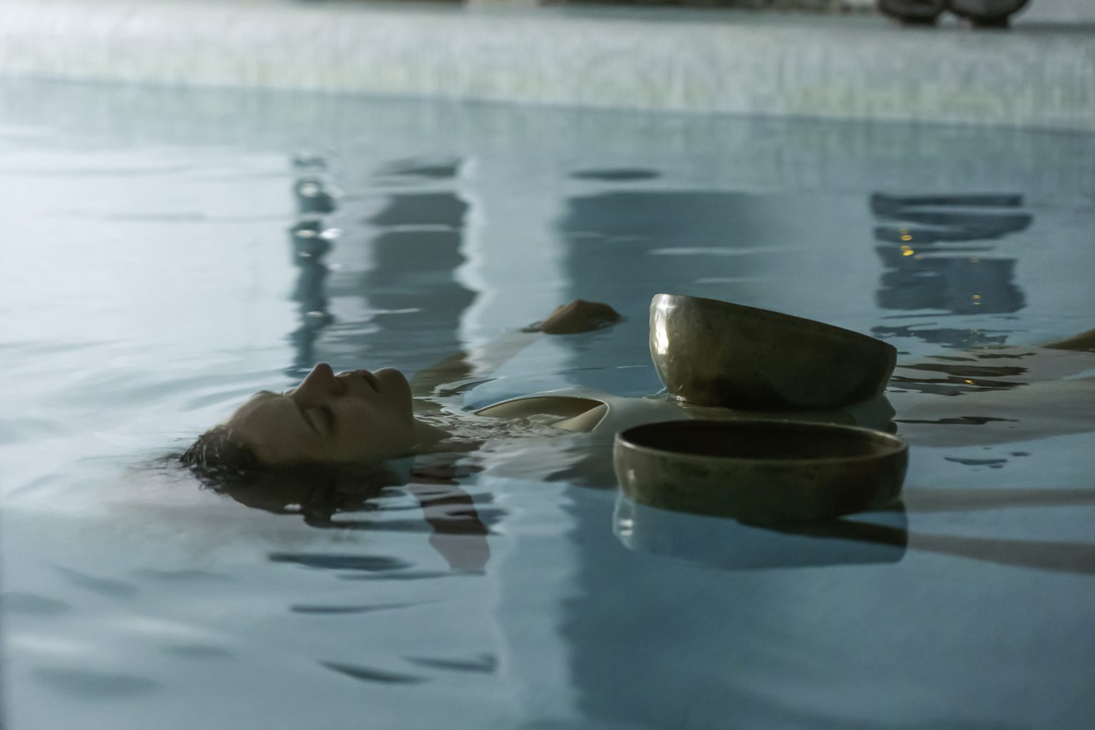 A guest floating in the thalassotherapy pool during a sound healing session, two singing bowls resting on the water