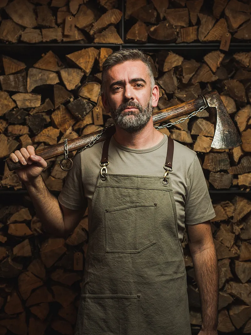 Chef Alexandre Silva with an axe against a stacked woodpile