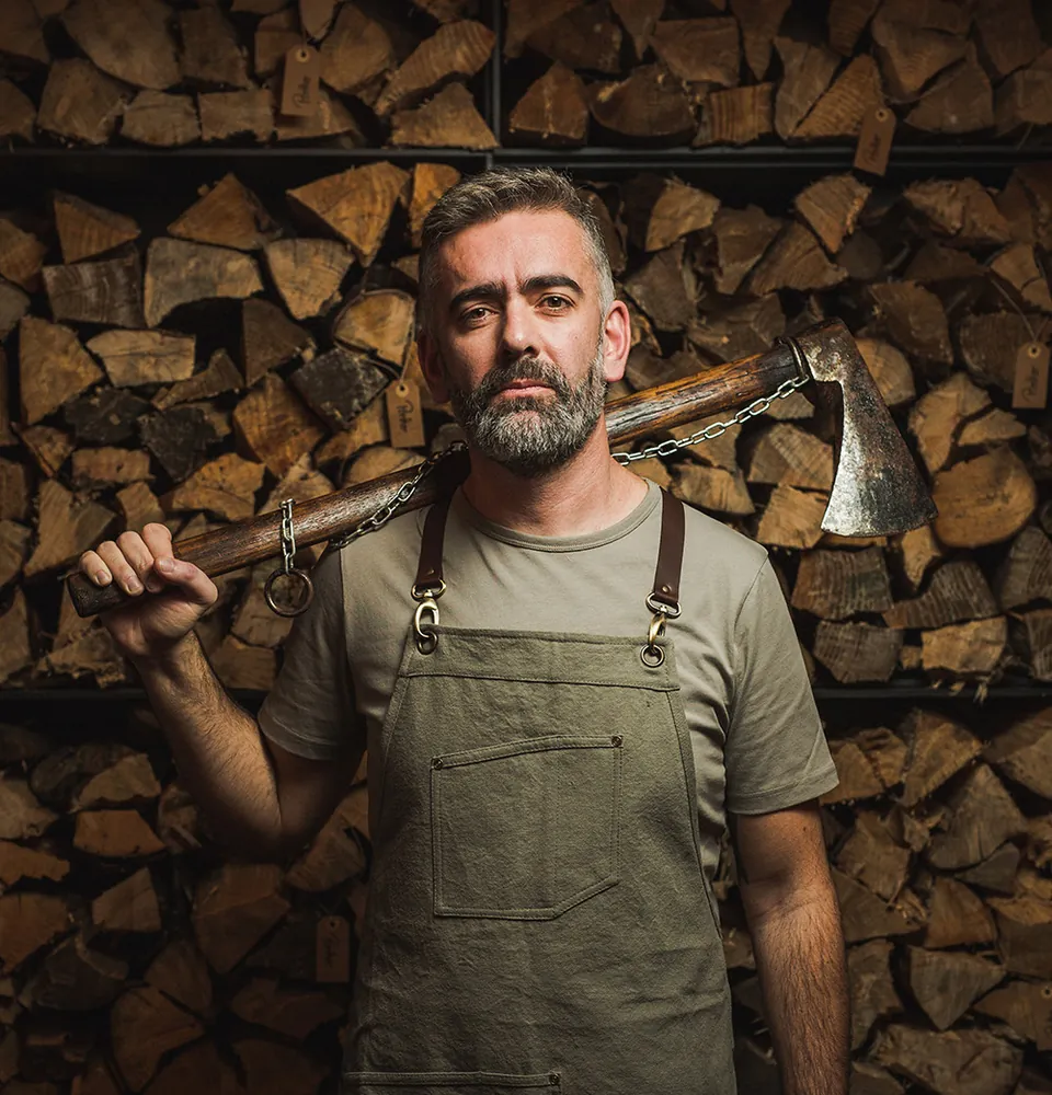 Chef Alexandre Silva with an axe against a stacked woodpile