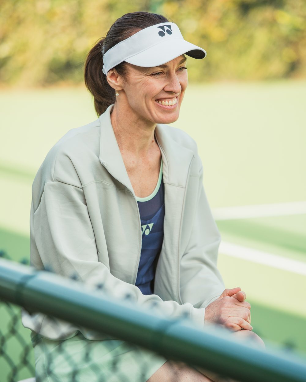 Martina Hingis smiling on court, in a pale green jacket and a white visor