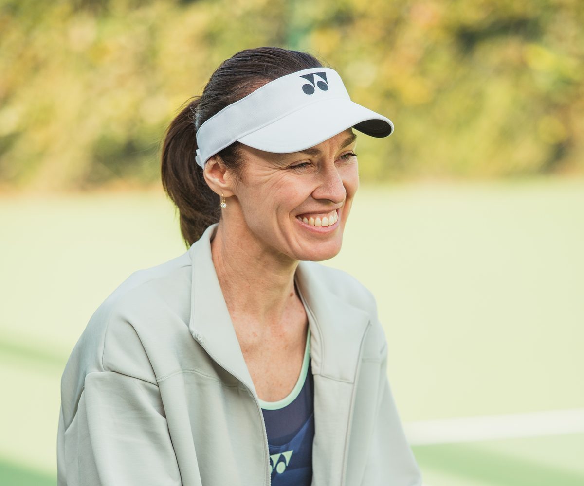 Martina Hingis smiling on court, in a pale green jacket and a white visor